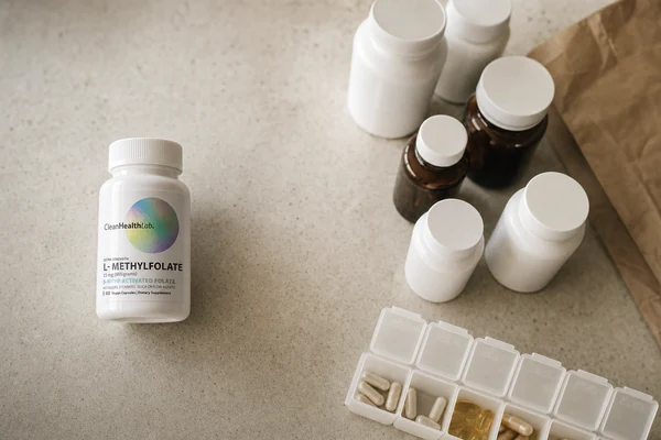 An overhead view of a kitchen counter: a cluster of unlabelled supplement bottles and a weekly pill organiser to one side, and a single Clean Health Lab L-Methylfolate bottle standing apart in focus.
