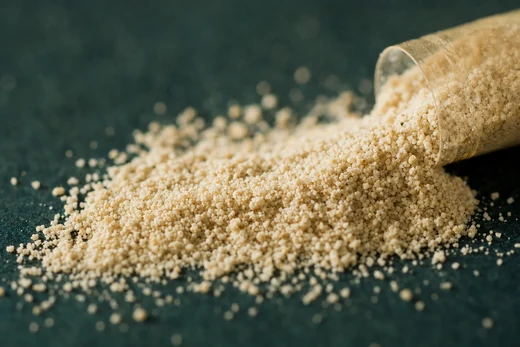 A macro photograph of pale crystalline folate powder spilling from a tipped measure onto a dark surface.
