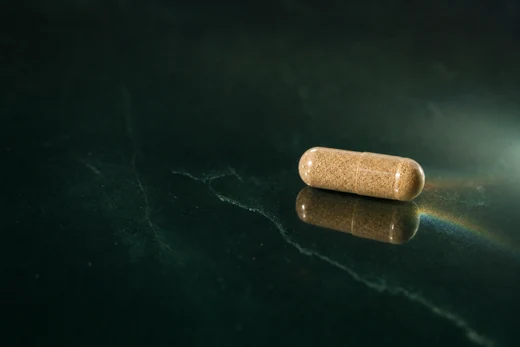One small tan capsule resting on polished dark jade, lit so the granular fill glows warm through the shell, with a clean reflection beneath it.