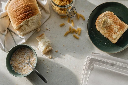 An overhead view of an ordinary breakfast on a limestone counter: a torn white loaf, a bowl of wheat cereal, dry pasta, buttered toast.