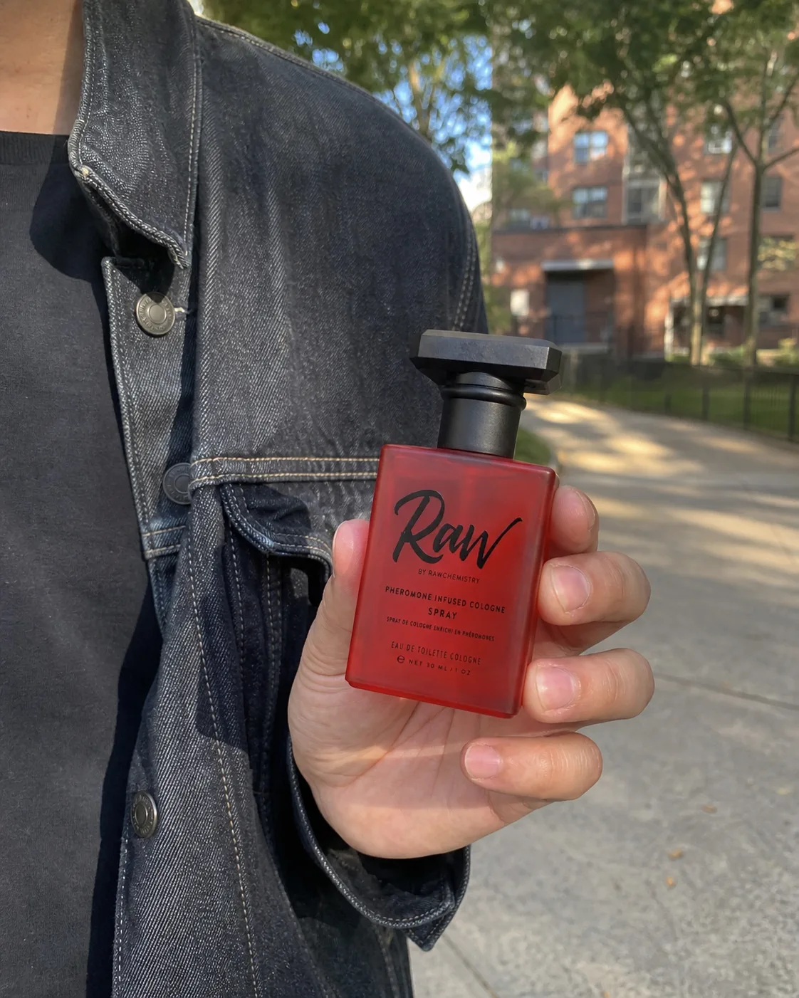 Man holding a RAW cologne bottle outdoors beside a denim jacket